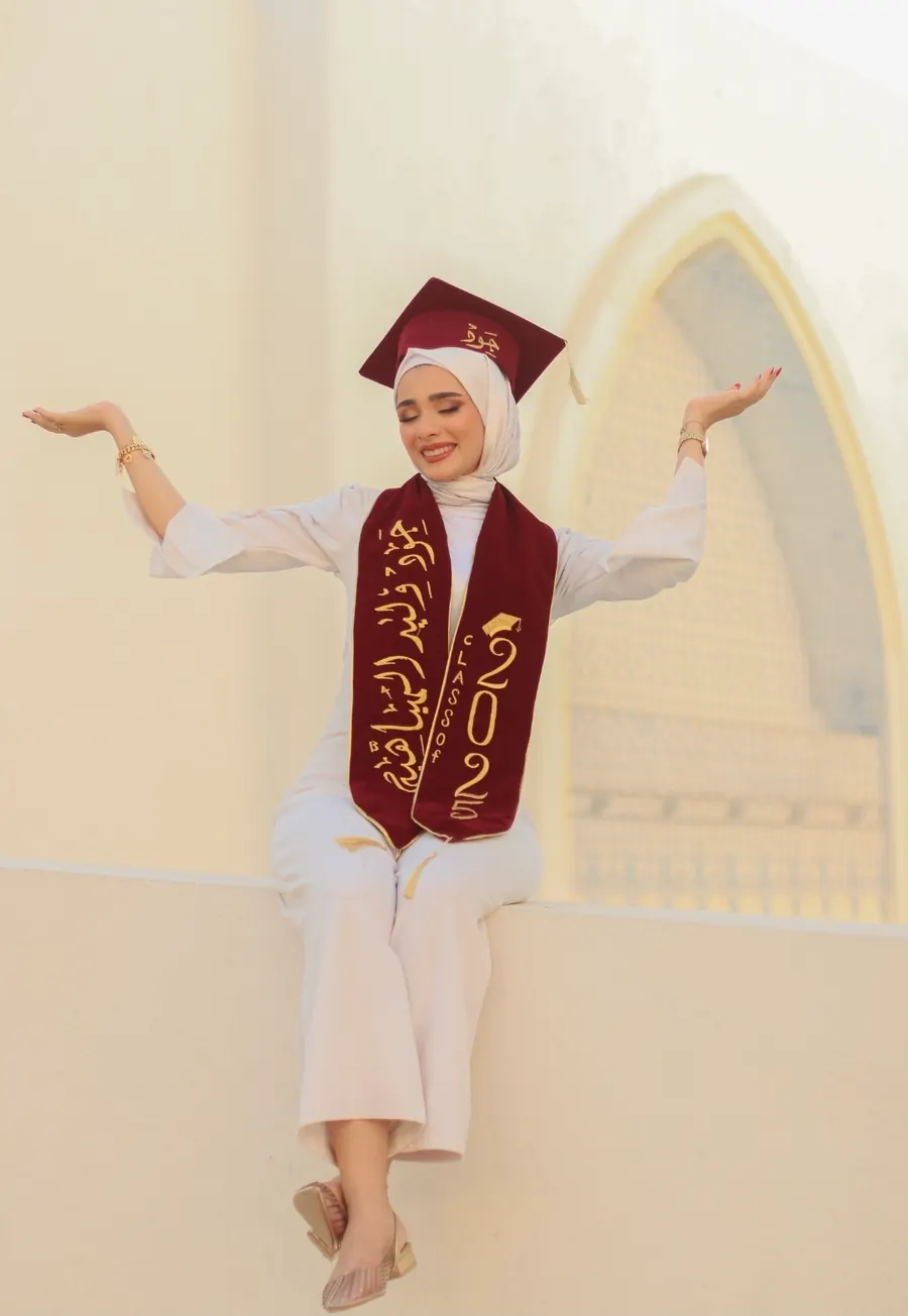 خرّيجة بقبّعة تخرّج وشال مطرّز تجلس على حافّة جدار وتفتح ذراعيها أمام نافذة مقوّسة