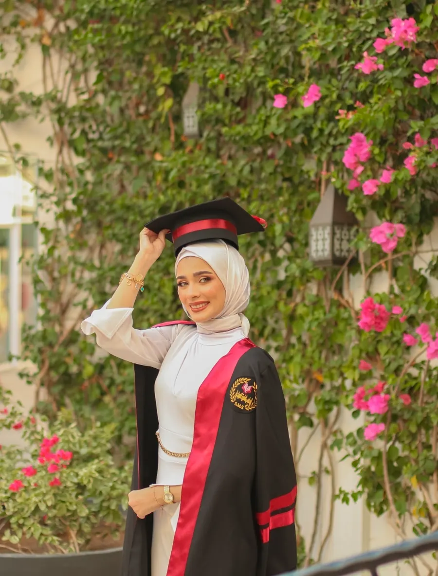 خرّيجة تعدّل قبّعة التخرّج بيدها أمام شجرة جهنمية وردية مزهرة