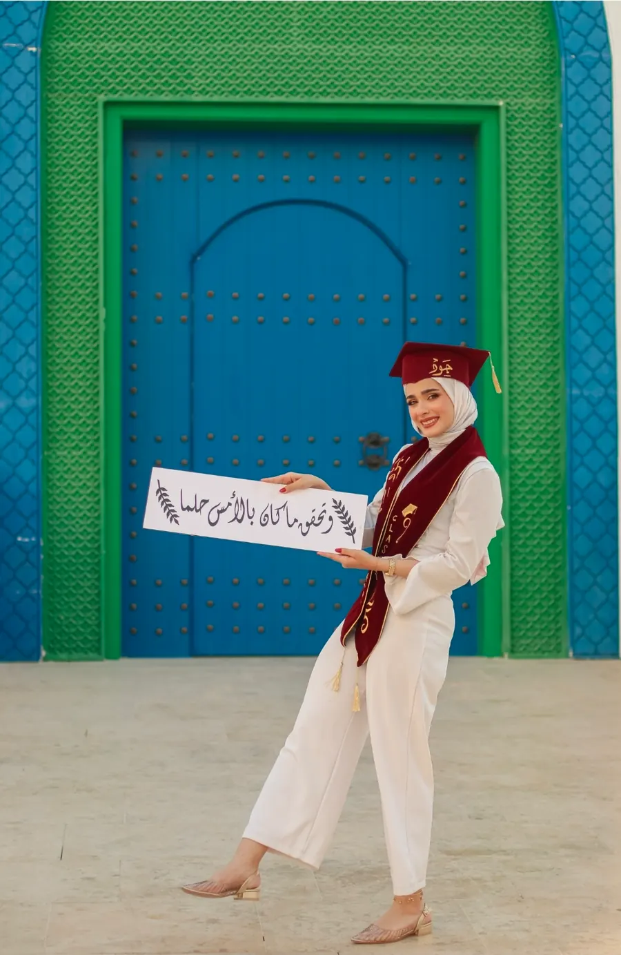 خرّيجة تحمل لافتة تخرّج مكتوبة بخطّ عربي أمام باب أزرق بإطار أخضر