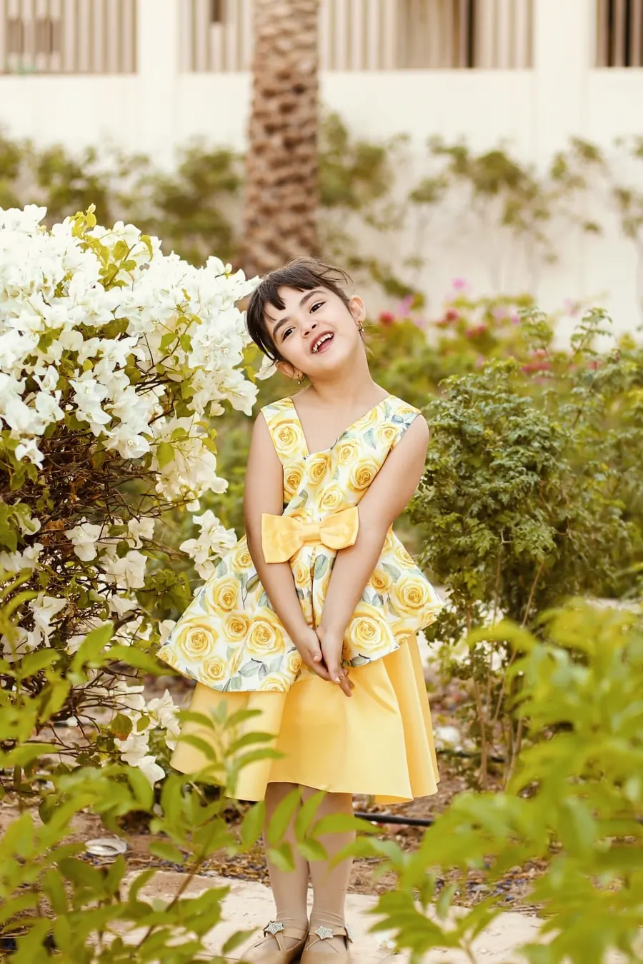 طفلة تضحك وتميل برأسها وتشبك يديها وسط نباتات خضراء وأزهار بيضاء